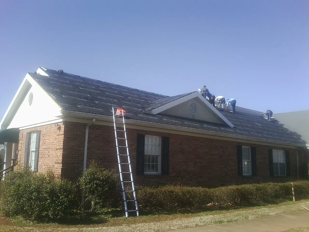 Crew tearing shingles off a brick ranch roof with a ladder set — Metal Roof Repair work by our crew in Wildersville