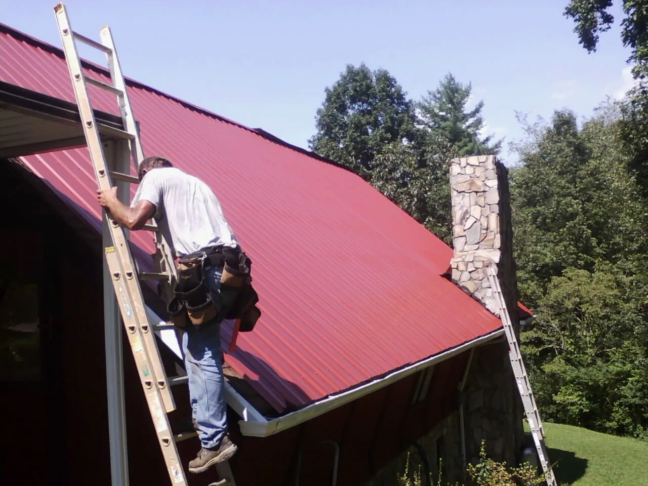 Roofer on a ladder beside a red metal roof and stone chimney — Soffit & Fascia Repair by Roofing Brothers in Wildersville